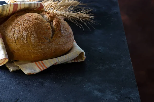 暗い背景に小麦の耳とナプキンと丸焼き素朴なライ麦丸パン、コピースペース — ストック写真