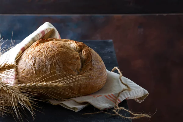 Redondo pan redondo de centeno rústico recién horneado con espigas de trigo y servilleta sobre un fondo oscuro, espacio para copiar —  Fotos de Stock