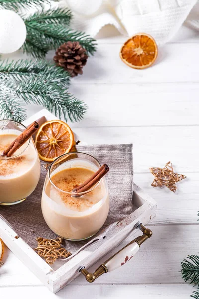 Зимний пряный горячий напиток эггног в стаканах с синамоном в лотке, сухой апельсин, елки и шишка на белом деревянном столе — стоковое фото