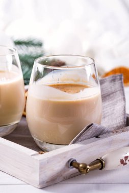 Baharatlı sıcak içecek, bardakta yumurta likörü tepside sinamon, beyaz ahşap masada köknar dalları.