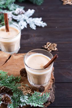 Tarçınlı eggnog Noel sütü kokteyli, iki bardakta sunulur bir tahta kesme tahtasında köknar dalı ve koyu ahşap arka planda çam kozalağı ile birlikte.