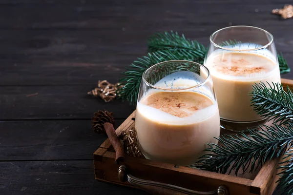 Tarçınlı eggnog Noel sütü kokteyli. Koyu ahşap arka planda köknar dalı olan antika bir tepside iki bardakta servis ediliyor.
