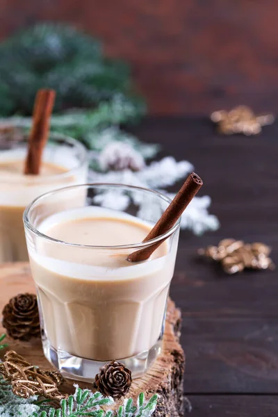 Cocktail de lait de Noël au lait de poule avec cannelle, servi dans deux verres sur une planche à découper en bois avec branche de sapin et cône de pin sur fond bois foncé — Photo