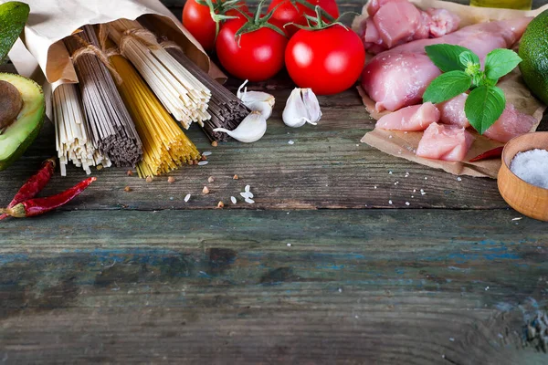 Bunch of Italian spaghetti, noodles soba and sommel, chicken raw meat, avocado with tomato on an old wooden background — Stock Photo, Image