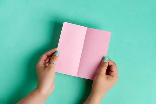 Mãos femininas estão segurando vazio brochura mock-up para escrever carta acima fundo azul-esverdeado pastel — Fotografia de Stock