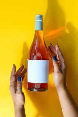 Havada ve gölgelerde sarı bir zemin üzerinde logo olan kadın ellerinde bir şişe gül şarabı. Üst görünüm.