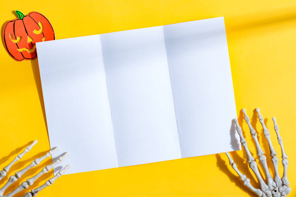 Пустой макет белой бумаги тройной брошюры A4 буклет в руках скелета на желтом фоне с ручной вырезанной тыквой, копировальное место. Вид сверху. Концепция Helloween.