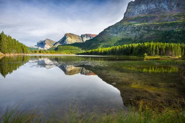 Kırmızı kayalar, çam ağaçları ve Montana, Buzul Ulusal Parkı 'ndaki Red Rock Gölü' ndeki temiz yansıtıcı su yaz öğleden sonraları