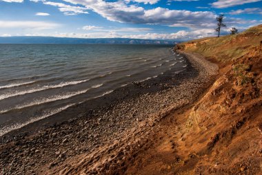 Arka planda dağlar ve güzel bulutlar ve ışık ile Baykal göl kenarında. Gölün yanında taşlar var. Sisteki dağlar.