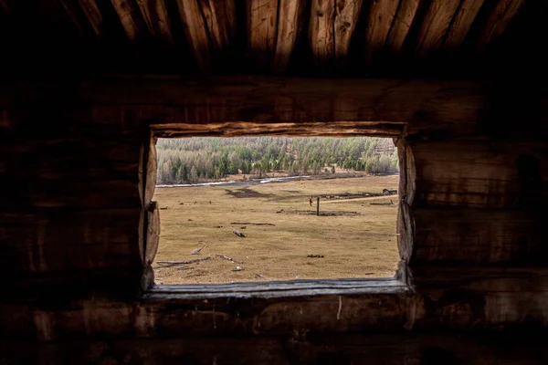 View from the log window to the field with a farm and a river. Frame of logs. Against the background of the forest, on the field of old logs. Brown and yellow tones.