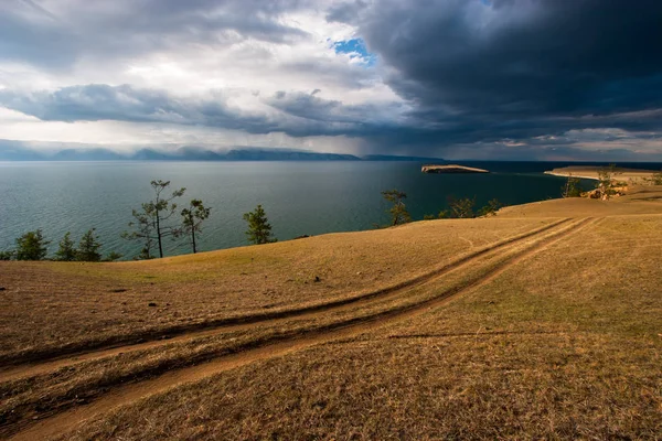 Steppe road on the shore of Lake Baikal. In the lake is an island. Rain clouds in the sky. Behind the lake are mountains over which it rains. Shades of yellow and blue.