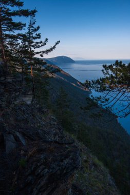 Akşam saatlerinde doğu yakasından Baykal Gölü kıyısındaki yüksek uçurum. Gökyüzü bulutsuz mavi. Kaya ağaçlarında.