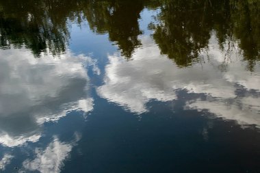 Nehrin suyundaki bulutların ve ağaçların yansıması. Gökyüzü mavi. Bulutlar beyazdır. Ağaçlar yeşildir. Su opak, yansıtır.