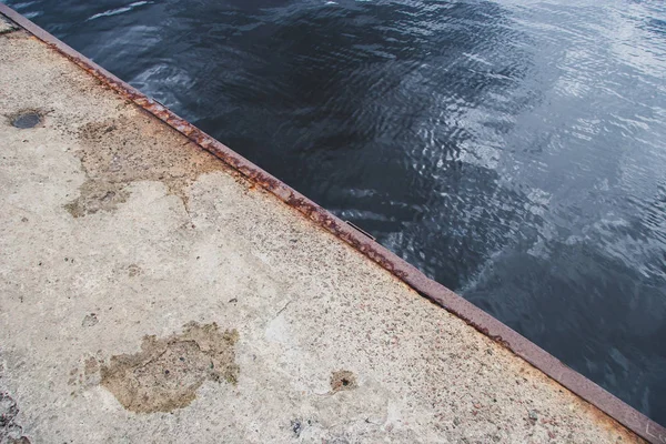 The edge of the old concrete pier with a rusty iron edging. Diagonal in the frame. Blue water with ripples and a reflection of the sky.