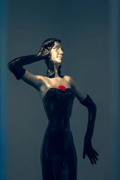 Woman\'s mannequin behind the glass with black hair in a black dress with a red rose on the dress and in long black gloves. Light blue and yellow. An interesting pose. One hand holds his head, the other hand is set aside.