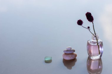 Minimalizm. Bir bardak, üç taştan oluşan bir piramit ve gül yağı ve sıkışmış bir dalile şişeler. Çakıl ve cam üzerinde seçici odak. Bulanık mavi arka plan. Metin için bir yer var.