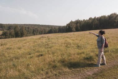 Bir kız turist ormanlık bir alanda bir şey gösterir. Çimenlerin ve çiçeklerin etrafında. Turist pantolonu giymiş kız, çizgili bir ceket ve kırmızı bir sırt çantası..