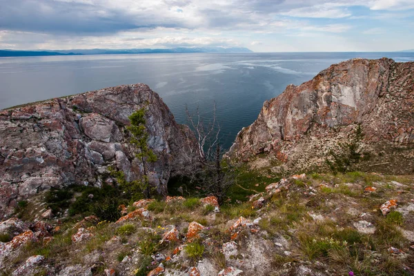 View between two rocks on Lake Baikal from Olkhon Island. On the stones is red moss, near the vets and grass. Mountains behind the lake. Clouds in the sky. There is a dry tree and green.