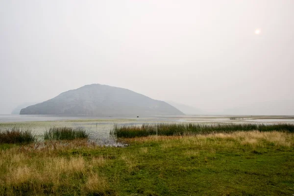 Lake in the fog and a fisherman in a boat with green grass near the shore. Peace and silence. Big rock in the water. The faded sun shines through the fog. Grey sky. Copy space.