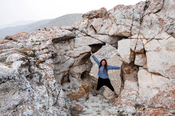 A smiling girl tourist stands in a natural stone arch with arms and legs spread apart like a star. Around the mountain. Cracked large stones with red moss.