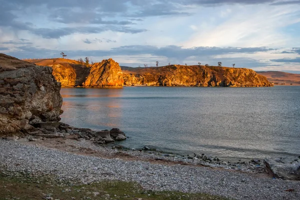 Gün batımında güneşin aydınlattığı göldeki kırmızı kayalar. Çakıl plajı. Gökyüzündeki bulutlar. Su sakin, dalgalar la ama dalgalar olmadan.