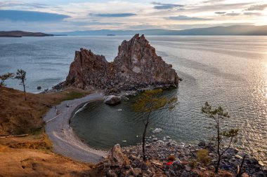 Pelerin üzerinde duran granit kayanın manzarası. Taşların ve çimenlerin kıyısında. Göl suyundaki dalgalar. Gölün ötesindeki dağlar. Günbatımı. Gökyüzünde bulutlar var..