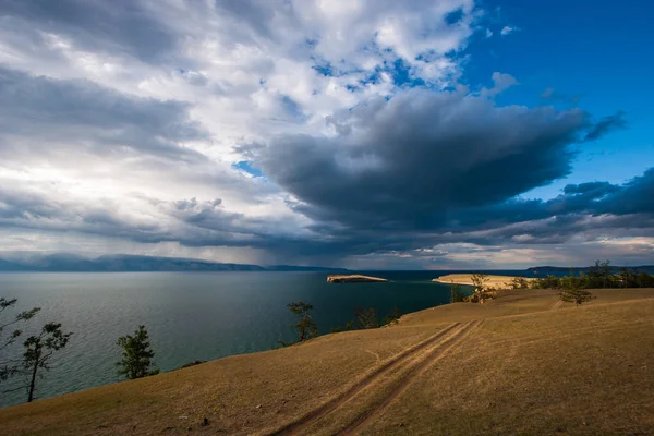 View of the island on Lake Baikal across the steppe with a field road in cloudy weather with the sun and a very beautiful sky with clouds. More Trees grow on the edge of the shore.