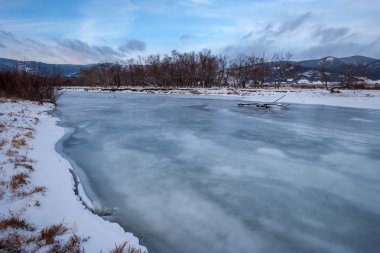 Buzlu kış nehri ve donmuş ağaçlar. Ufukta tepe var. Kıyıda kar ve ağaçlar büyür..