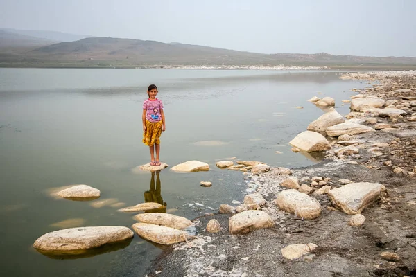 A child girl in a striped T-shirt and a yellow skirt is standing on a stone in the water in the lake. Misty sky and hills from behind. A lot of big white stones. Copy space.