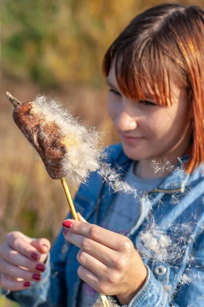 A child girl with red hair holds a cattail in her hand and watches the fluff fly around. The girl is smiling. Selective focus on the plant.