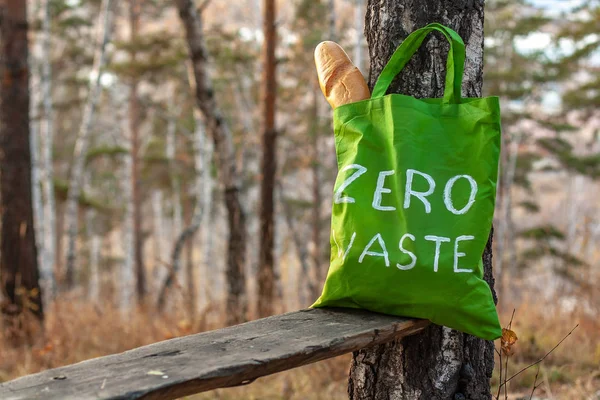 Textile green bag with the inscription zero waste on a background of nature. French bread in a bag. Selective focus. Blurry trees and autumn grass on the background.