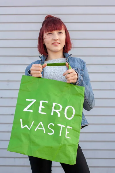 Green eco bag with a white inscription zero waste held by a young girl with red hair. Smiling girl in a denim jacket on a striped gray background.