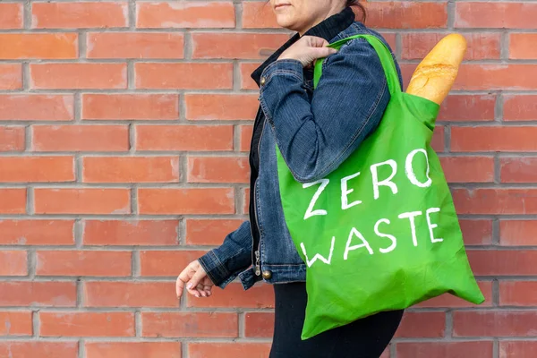 A girl holds a green textile eco bag with French bread and zero waste on her shoulder. Girl in a denim jacket on a background of a red brick wall with copy space.