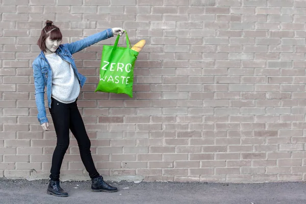 The girl is holding on her outstretched arm a textile eco bag with the inscription zero waste from which French bread sticks out. The brick wall is discolored. Copy space on the right.