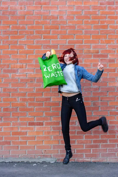 Smiling young girl in a jump with an eco bag with white text zero waste. The girl has red short hair, French bread is visible in the bag. Copy space. Vertical.