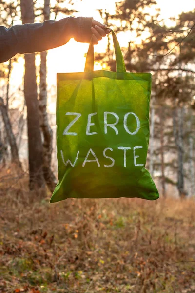 Zero waste concept. Textile green bag with a white inscription in hand on a background of nature and the sun. Against the background of blurry trees and autumn grass. Vertical. Copy space below.