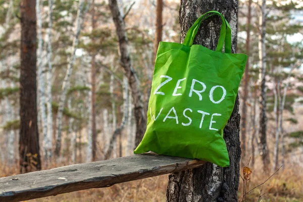 Standing in a forest on a bench near a birch, a textile green bag with the inscription zero waste. Selective focus. Blurry trees and autumn grass on the background.