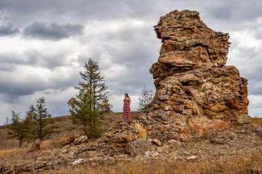 Bir kız gökyüzüne karşı taşın yanında duruyor. Harap olmuş eski taşlar. Yakınlarda birkaç ağaç var. Gökyüzündeki bulutlar. Yatay.