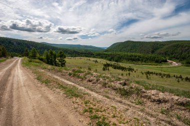 Yeşil dağlarla orman arasında dolambaçlı bir yol var. Toprak yol. Mavi gökyüzünde bulutlar. Güneşli. Yatay.