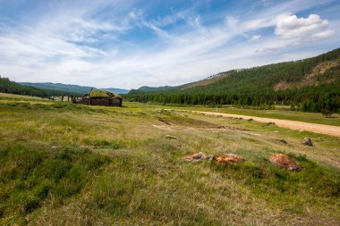 Tepeler arasında toprak bir yol ve çimlerin üzerinde harap olmuş eski bir yurt. Çimen kulübesi ile kaplanmış. Mavi gökyüzünde bulutlar. Güneşli. Yatay.