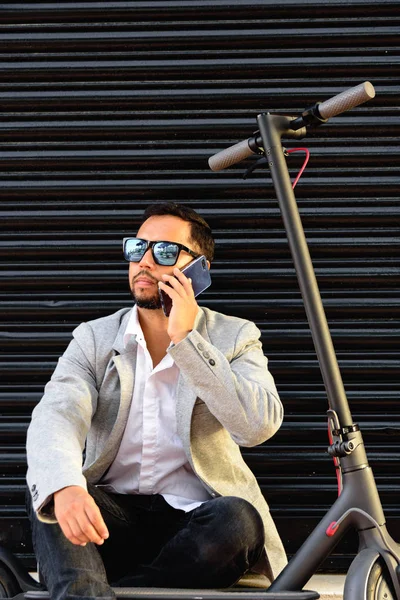 Latin adult man with sunglasses, well dressed and electric scooter talking on his mobile phone sitting on the street with a black blind in the background