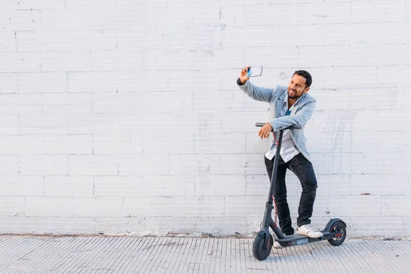 サングラスをかけたラテン系の大人の男性、よく服を着て電動スクーターは、白い壁の背景を持つ通りで彼の携帯電話で自分撮りを取ります — ストック写真