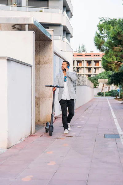 Lateinischer erwachsener Mann mit Sonnenbrille, gut gekleidet und Elektroroller, der auf der Straße mit seinem Handy spricht — Stockfoto