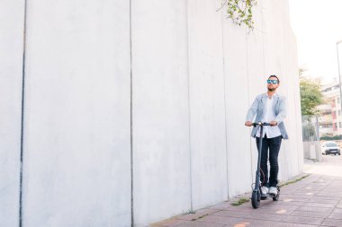 Güneş gözlüğü ile Latin yetişkin adam, iyi giyimli ve beyaz kör arka plan ile sokakta elektrikli scooter sürücü