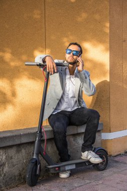 Güneş gözlüğü ile Latin yetişkin adam, iyi giyimli ve elektrikli scooter arka planda siyah bir kör ile sokakta oturan cep telefonu konuşuyor