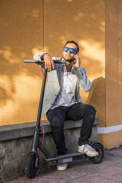 Homem adulto latino com óculos de sol, bem vestido e scooter elétrico falando em seu telefone celular sentado na rua com uma cega preta no fundo — Fotografia de Stock