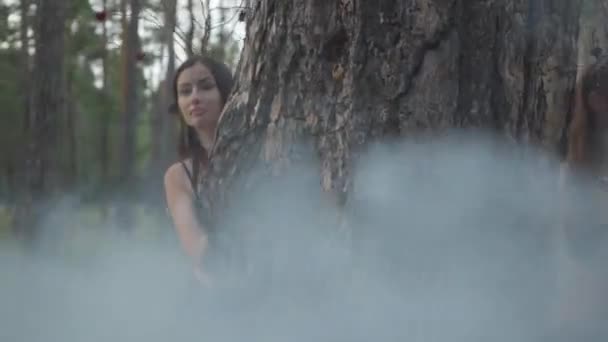Привлекательные дриады или лесные феи выходят из-за ствола дерева и танцуют в красивых костюмах в облаке дыма. Древний ритуал лесных созданий. Выступление танцоров в лесу — стоковое видео