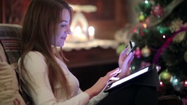 Beyaz kadın Noel ağacı yakınında koltukta oturan ve kredi kartı ile online alışveriş yaparken dijital tablet kullanarak video 