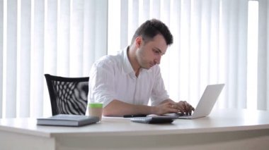 Genç adam ofiste oturuyor ve dizüstü bilgisayarda yazı yazıyor. Bir kalem al ve kağıda yazmaya başla. Adam kameraya el sallıyor..