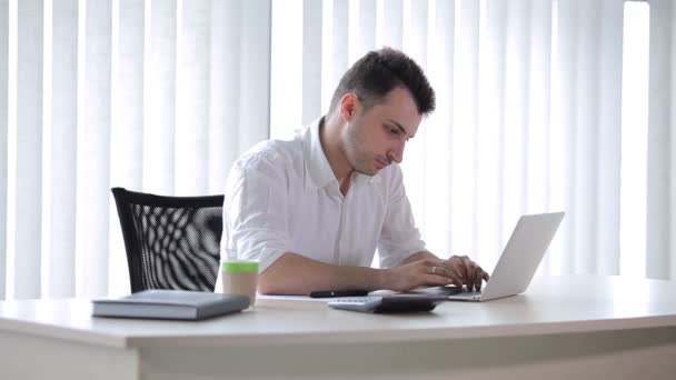 这个年轻人坐在办公室里 在笔记本电脑上打字 人类拿起一支笔 开始在纸上写字 男人在镜头前挥手 — 图库视频影像